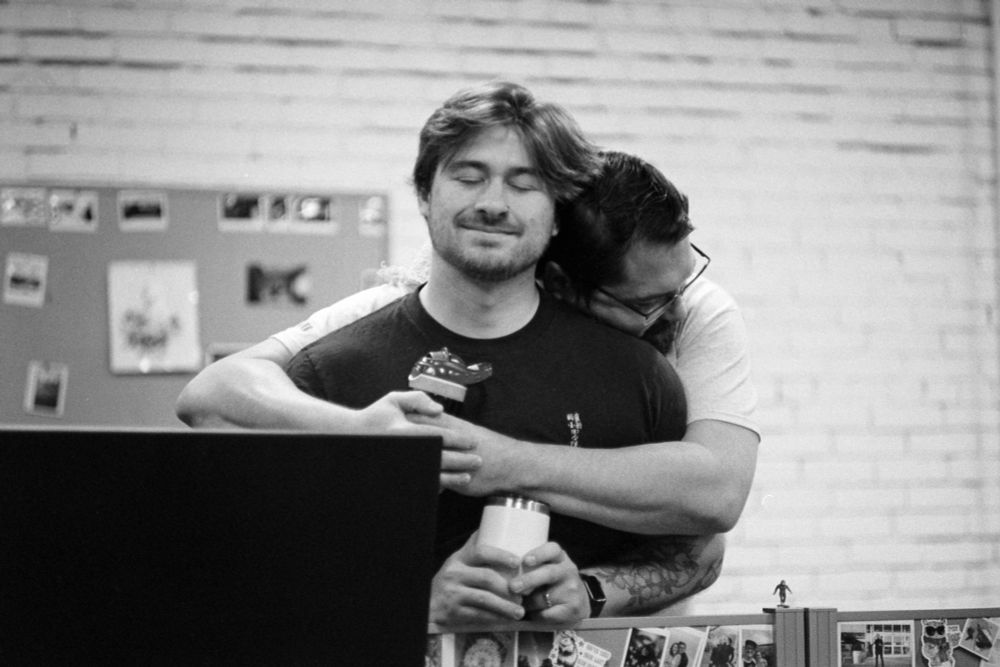 A black-and-white photo of a man smiling as another man hugs him.