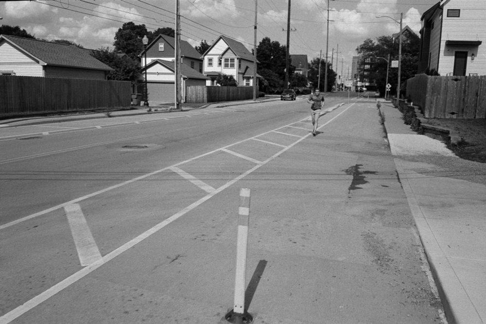A black-and-white photo of a man jogging in a divided bike lane as if it's a multi-use path, right beside a clear sidewalk.