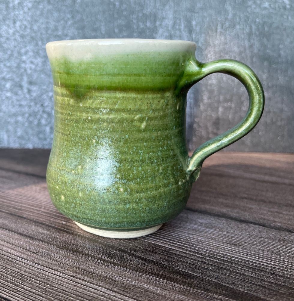 Green mug with handle to the right hand side. The body of the mug sits in a small unglazed foot. The form bellies out at the base. The middle of the mug curves inward slightly. The rim has a clear glaze, revealing the white clay body.

Laguna Bmix with grog, Laguna fern mist green, clear liner glaze. Cone 6.