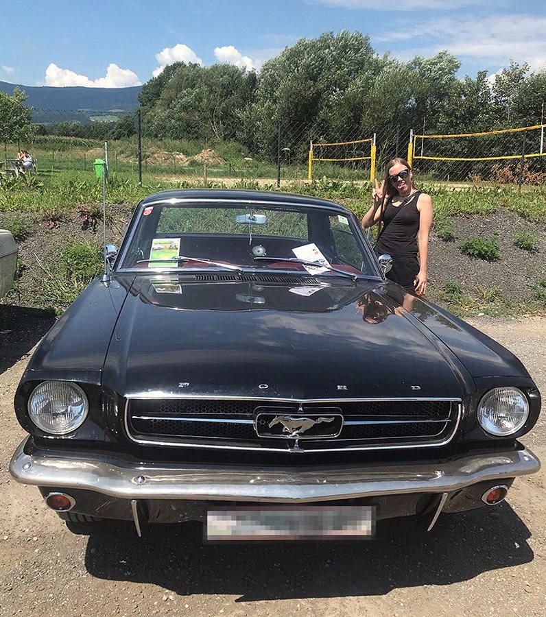 a photo of a black ford mustang car from the front. it has some paper on the windshield, the leather inside is red and it stands on a dirt road with gras and trees in the background. Also there is a silly goober standing next to it. 
