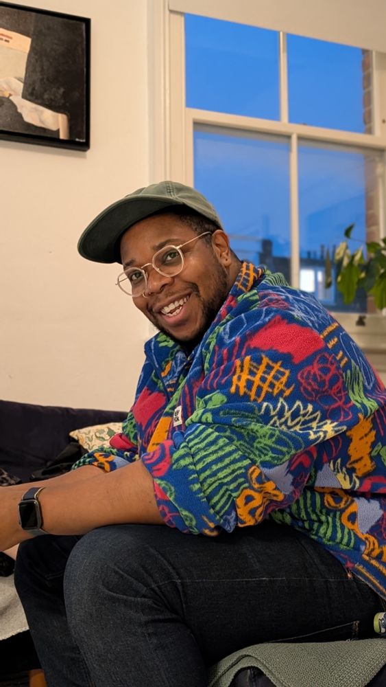 A black man in a multicoloured/multi patterned fleece, green hat and glasses. 
Smiling directly at the camera 