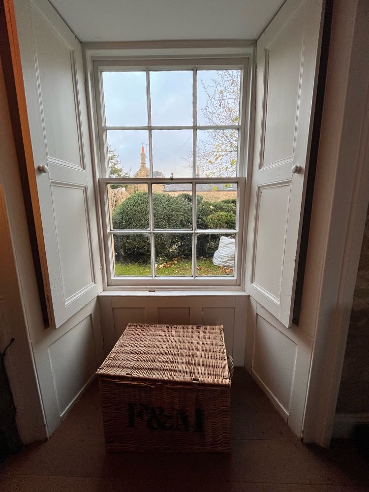A window leading outside with a hedge and old building in the background. 

A brown basket is just below the window in the foreground 