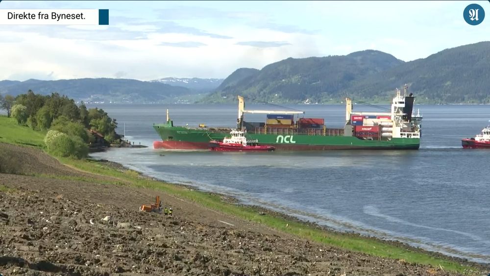 A large container ship reversing away from the shore of a fjord assisted by two tugs.