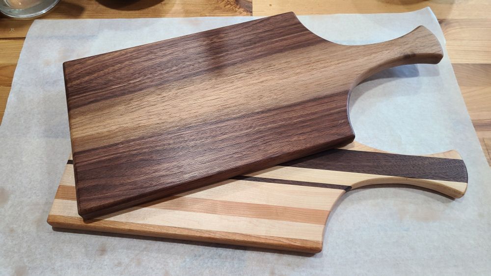 Two serving boards with handles, one on top of the other. The upper one is walnut and features a gorgeous grain pattern alternating between heart wood and sap wood. The lower one is made of strips of maple, cherry, and walnut.