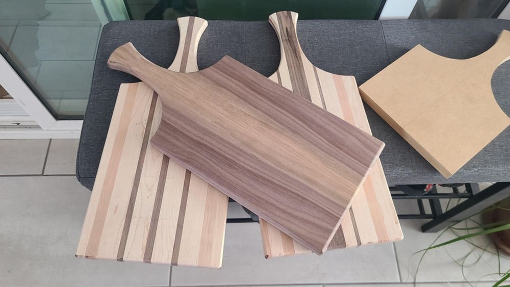 Three unfinished charcuterie boards with handles sit on a bench alongside a template for the handle.
