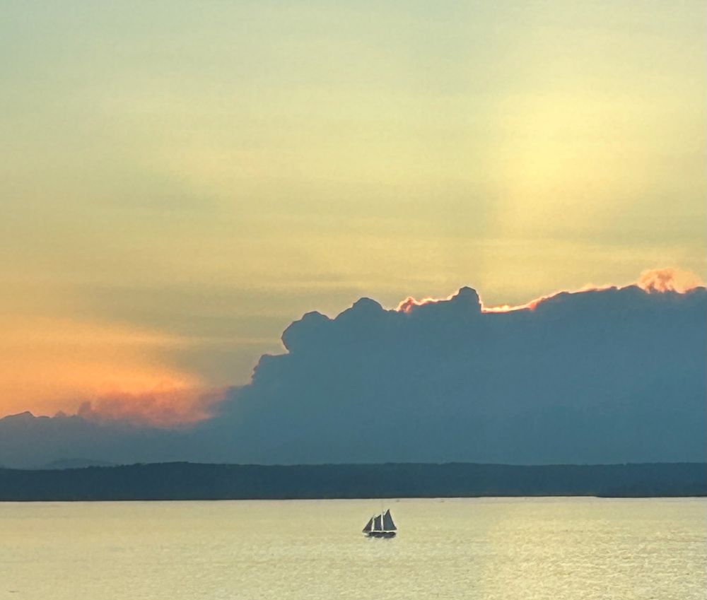 Photo from Seattle at (across Elliott Bay) of smoke building over the Olympics, and spreading north. A small sailboat on the water in the foreground. Sun has just set behind the column of smoke.