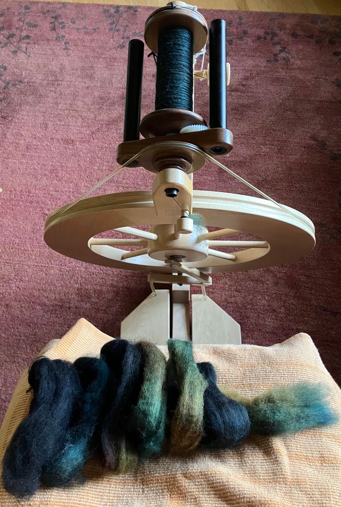An orange handwoven lap towel in the foreground ahead of a Lendrum upright spinning wheel. On top of the towel rests a small amount of combed wool top - a coil of about 6 loops (about the size to wrap around a hand, not coincidentally) in black, teal, chestnut, and a mossy green. 