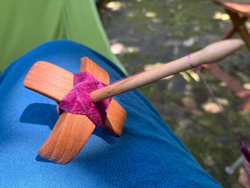 First time I thought about stopping today. The cop is magenta with faint shading towards purple on the outer wrapping layers. The spindle rests on a medium-blue pants leg with a bright green camping chair in the background giving very strobn color contrast. 