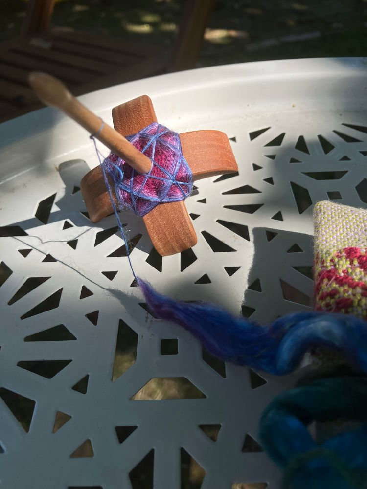 Third attempt to quit for the day. The spindle sits on the same gray table. The cop has an even wider purple band on the first layer of the cop and a second layer has been started. The braid and the little wrist-distaff cloth bag are in the shade in the foreground. 