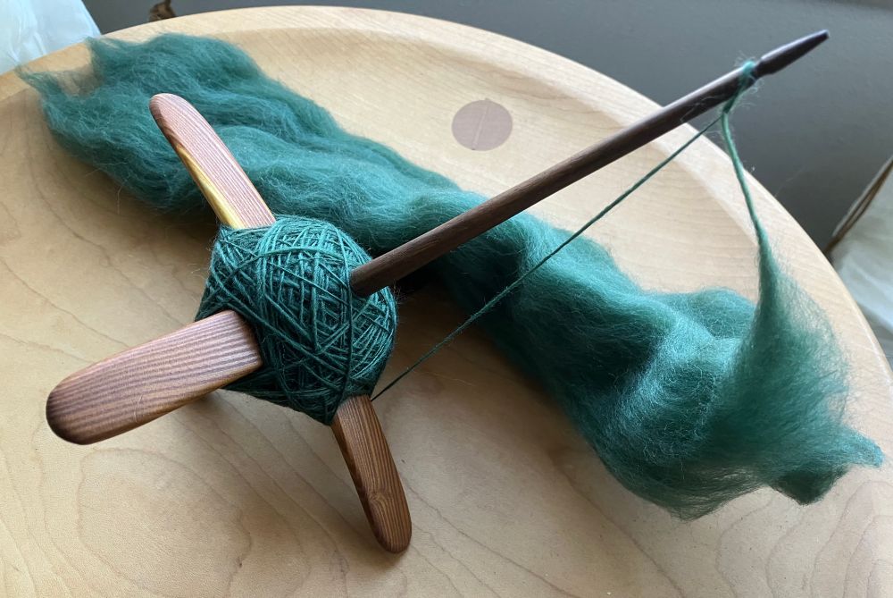 A cross-arm spindle with a blue-green cop of yarn and about a foot long strip of combed top. 