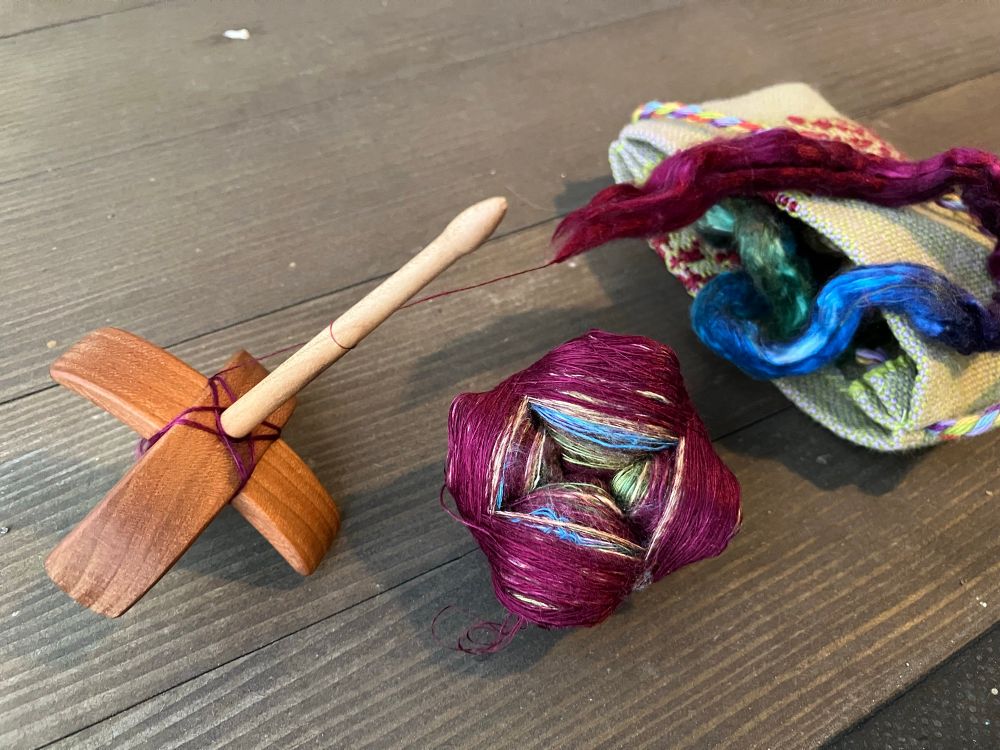 Back in the stage 13, a full cop removed from the small wooden spindle and resting beside it as a center-pull ball. The spindle has maybe a dozen wraps of very fine wine-colored silk thread on it - just enough to define the wrapping pattern. A small bag with the multicolored silk braids rests in the background. 
