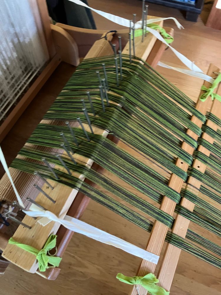 The warp and back beams of a small table loom. A homemade raffle (with nails as dividers) is lashed to the back beam and it is guiding the beaming of a two-toned warp. The warp is dark green and medium green 10/2 cotton. It is under tension and runs through lease sticks with a raddle cross a few inches away from the raddle. 