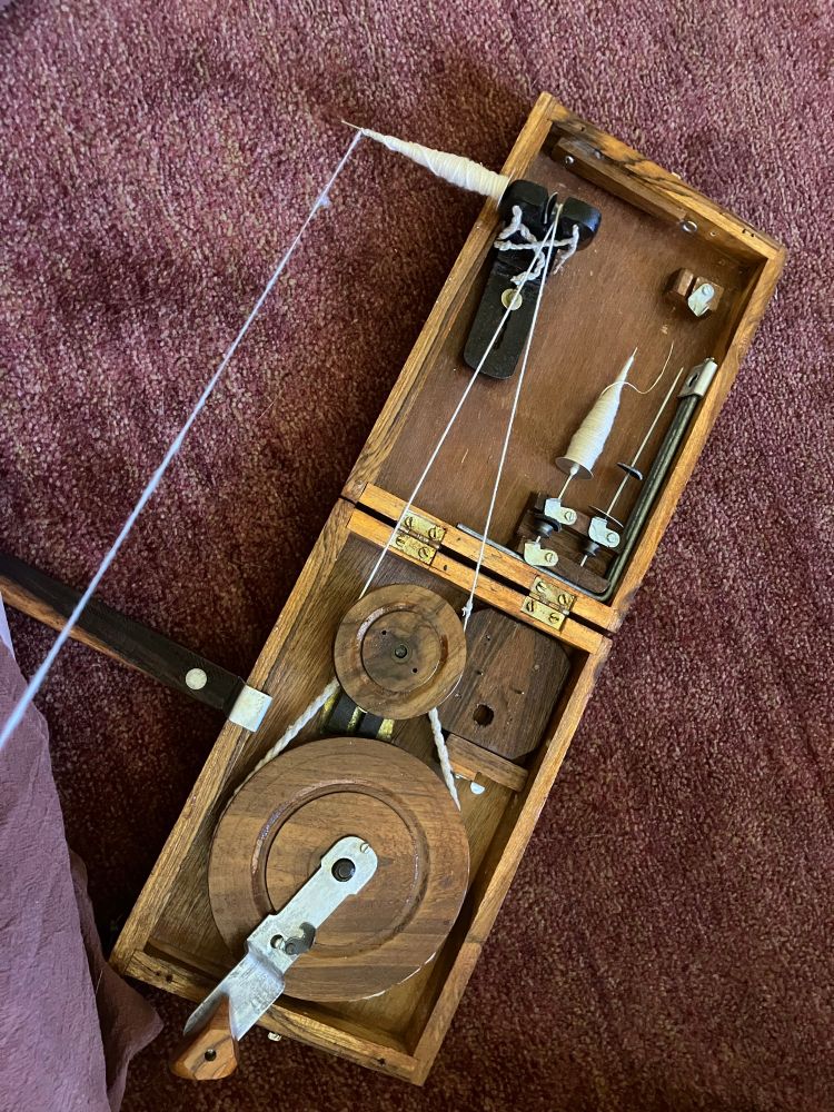 A book charkha spinning wheel from India. The “book” opens to have a large hand-cranked wheel that accelerates a smaller drive wheel that turns a metal wire spindle. Cotton yarn, not very evenly spun, is being wound on to the spindle. Another full spindle is stored inside the charkha box. It is very hard to take a picture of this process without it featuring mostly the foot that holds the box onto the ground to keep it from moving around. 