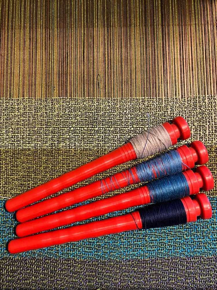 Four orange pirns resting on the web of a weaving-in-progress. The loom structure is not visible. The web has four stripes of weft colors each about two inches long. The stripes are dark blue, bright blue, yellowy-green, and a green so pale it is almost white. The pirns (an end-delivery counterpart of a bobbin) are each wound with very small amounts of thread. One has 40/2 unbleached linen, and the others have different shades of 20/2 blue mercerized cottons. 