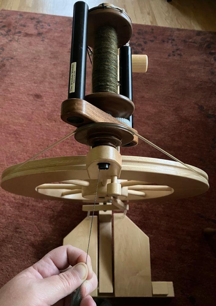 A Lendrum spinning wheel with a woolee winder flyer and a bobbin of fine yarn. The visible layer of yarn is a dark mossy green. The left hand of a white person (me!) is holding the yarn under tension. The fineness of the yarn is visible as it contrasts with the blond wood of the spinning wheel’s treadles. 