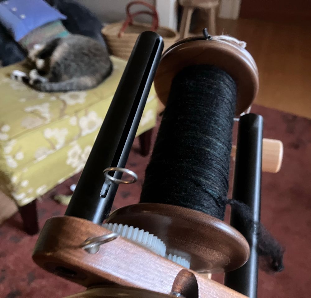 In the foreground, a walnut wood Woolee Windee flyer and a bobbin about two-thirds full of fine yarn. The outer layer of yarn is black but there are some under layers of teal and russet peeking through. In the background, a tabby cat with white paws is asleep on a footstool. Her white chin is aimed up at the ceiling but the back half of her body is curled up 