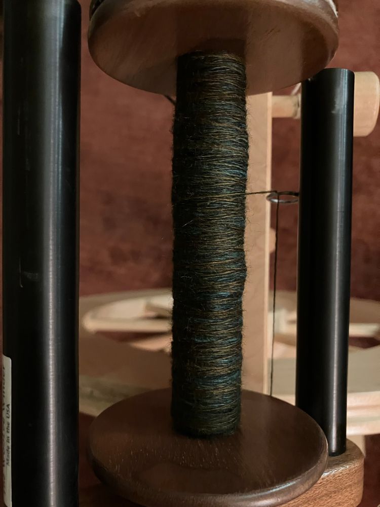 A zoomed in view of a bobbin and flyer. The yarn is fine and smooth, with dark cool jewel tones - black, dark teal, deep mossy green. 