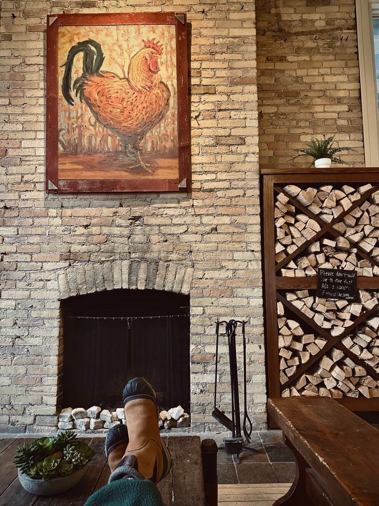 A rooster painting, stacked firewood and a cozy brick fireplace with two crossed feet in front of it. 
