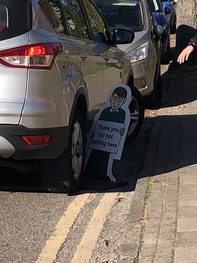A large silver car parked with a tyre on the foot of a sign showing a school boy holding a placard saying "Thank you for not parking here".