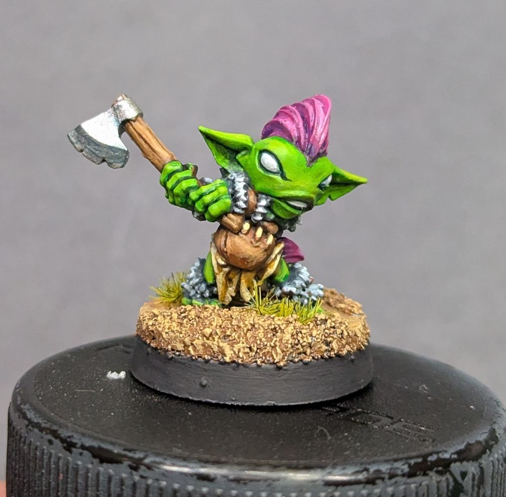 A small green goblin miniature resembling a classic fantasy Berserker - leather clothing with white fur trim, a purple Mohawk with a braid in the back, wildly swinging an axe.