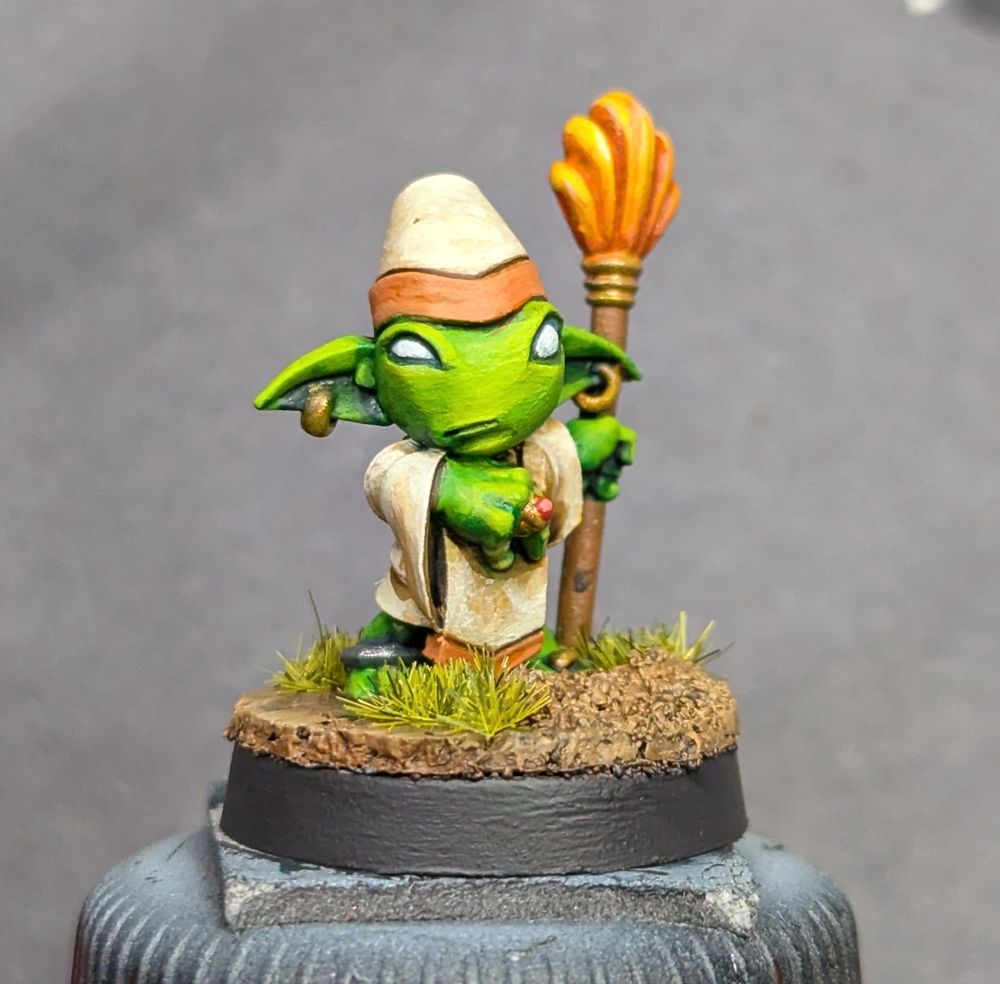 A small green goblin miniature, dressed as a sort of cleric, with earrings and a staff. He's wearing linen garments with orange trim.