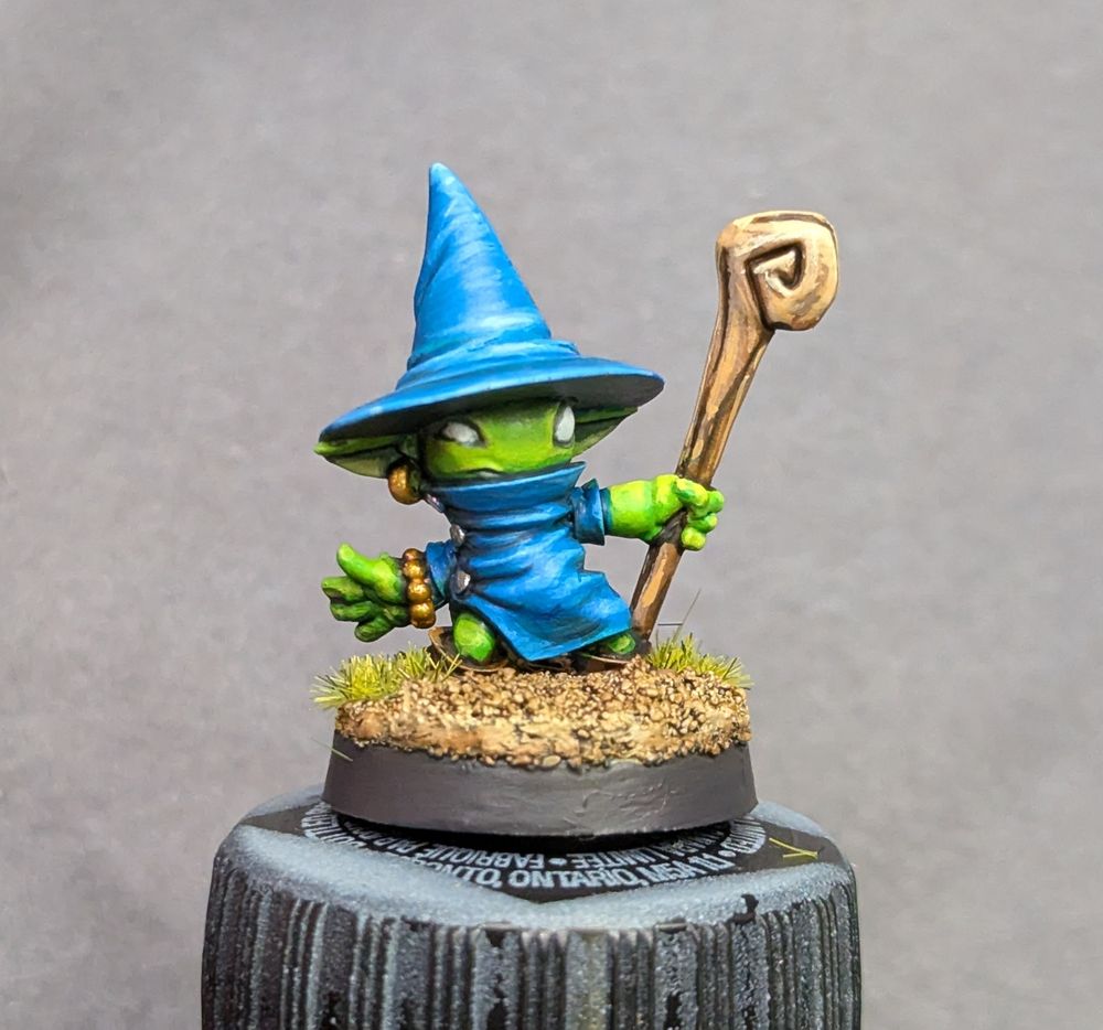 A small green goblin dressed in blue robes and a wizard hat, with a wooden staff.