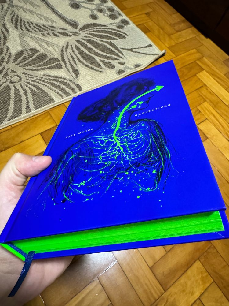 de um livro de capa dura azul com as laterais das páginas na cor verde limão. O título é “radioativas” da autora Kate moore. A ilustração na capa é a silhueta de uma mulher, desenhado com tinta preta, com detalhes em verde limão