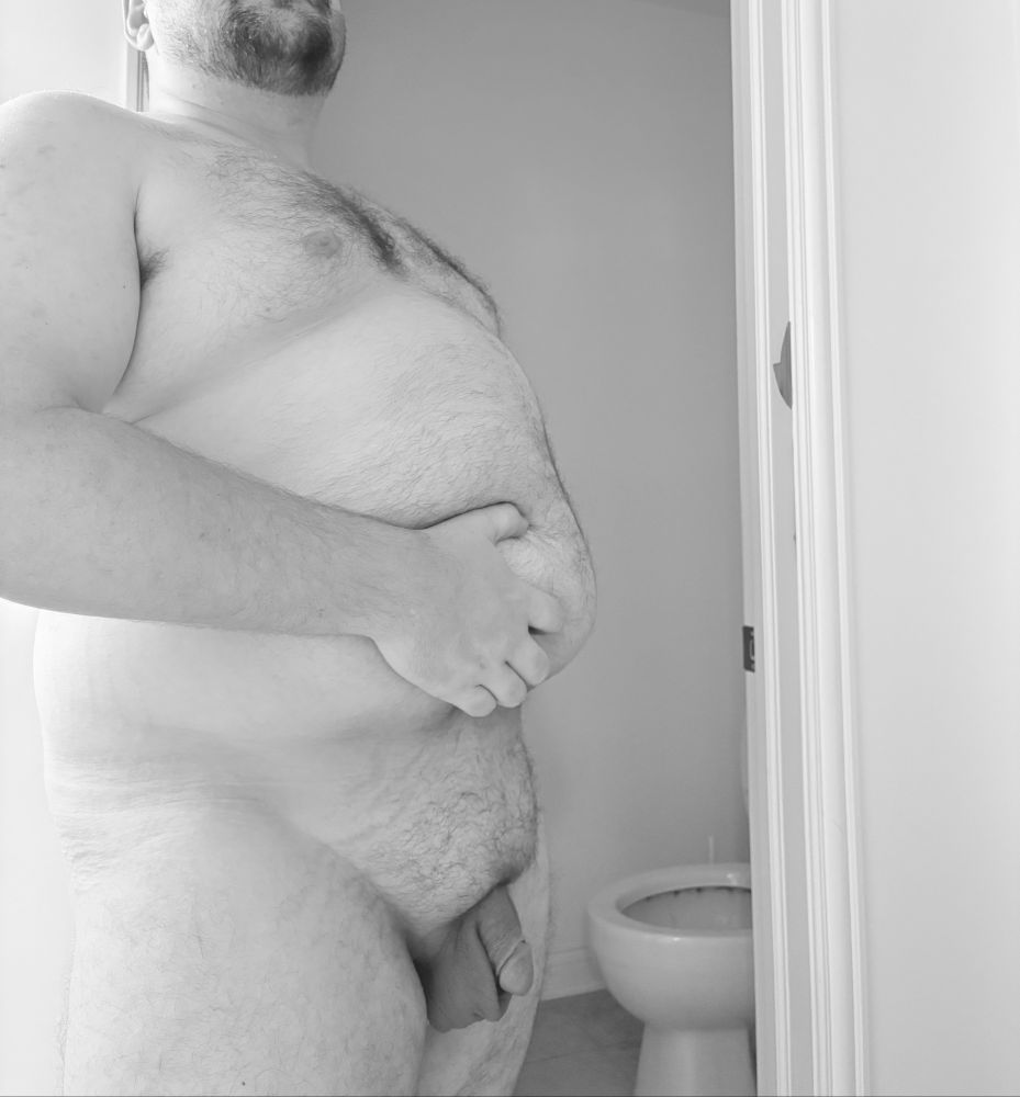 A black and white tasteful nude showing me standing naked, grabbing my bear belly, with a humorous toilet in the background