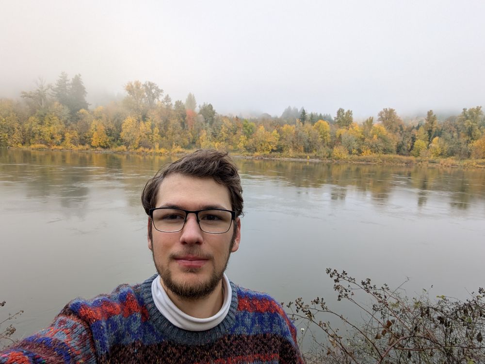 selfie of myself in front of a river lined with fall trees/colors
