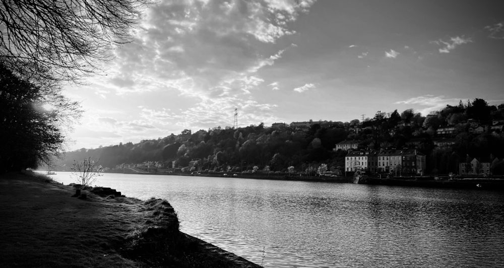 A black-and-white view from the Marina in Cork City, Ireland, looking across the river towards the Montenotte ridge. Scene of calm waters, tree-lined banks, and houses on a hillside, under a sky dotted with clouds.