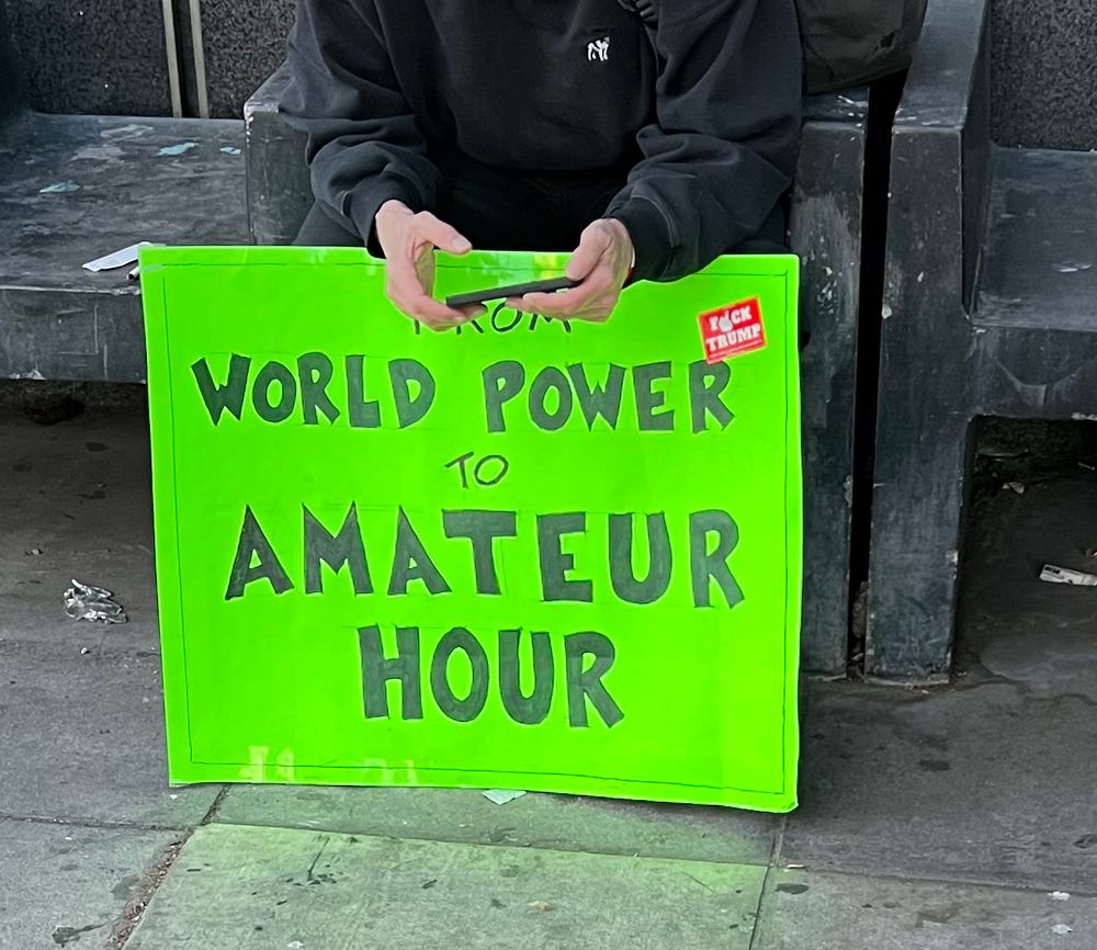 A man sits on a bench with a neon green sign with black lettering that says “FROM WORLD POWER TO AMATEUR HOUR” while taking a break at the Los Angeles chapter of the “Hands off” nationwide protests held in all 50 states and around the world in protest of Donald Trump, Elon Musk, DOGE, and the Republican Congress’ complacency in letting them gut our government and destroy our democracy.