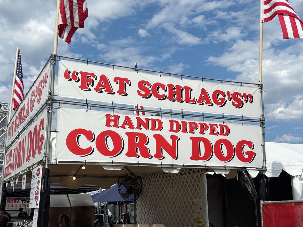 A sign on a state fair good stand that reads “Fat Schlag’s” Hand Dipped Corn Dog