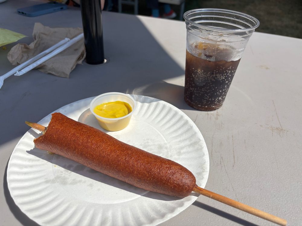 A jumbo corn dog on a paper plate with mustard and coke on a hot day at the state fair 