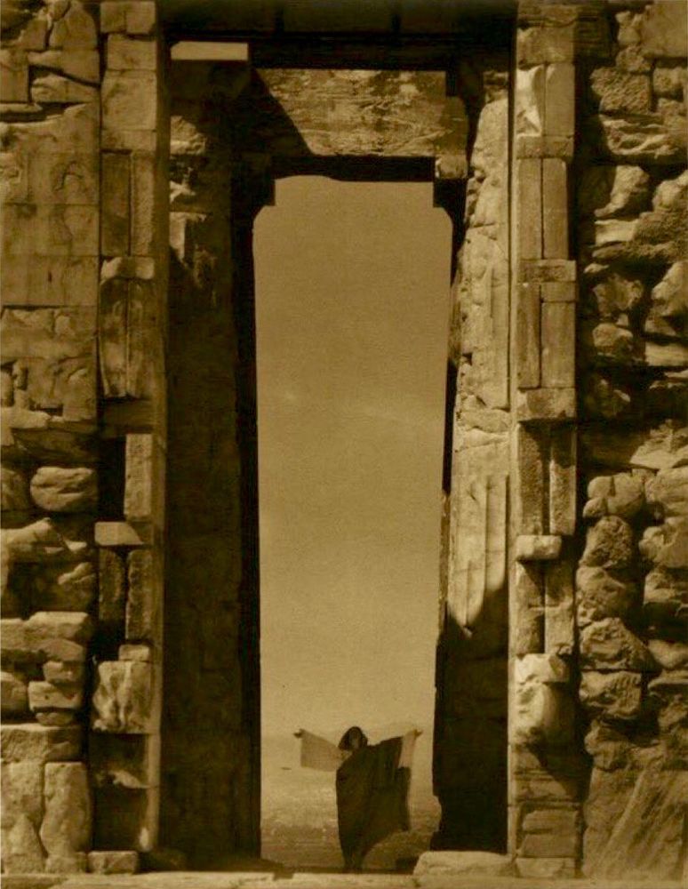Isadora Duncan at the Parthenon photographed by Edward Steichen: a woman in flowing gowns with long dark hair and arms outstretched stands between huge columns of the ancient structure.
