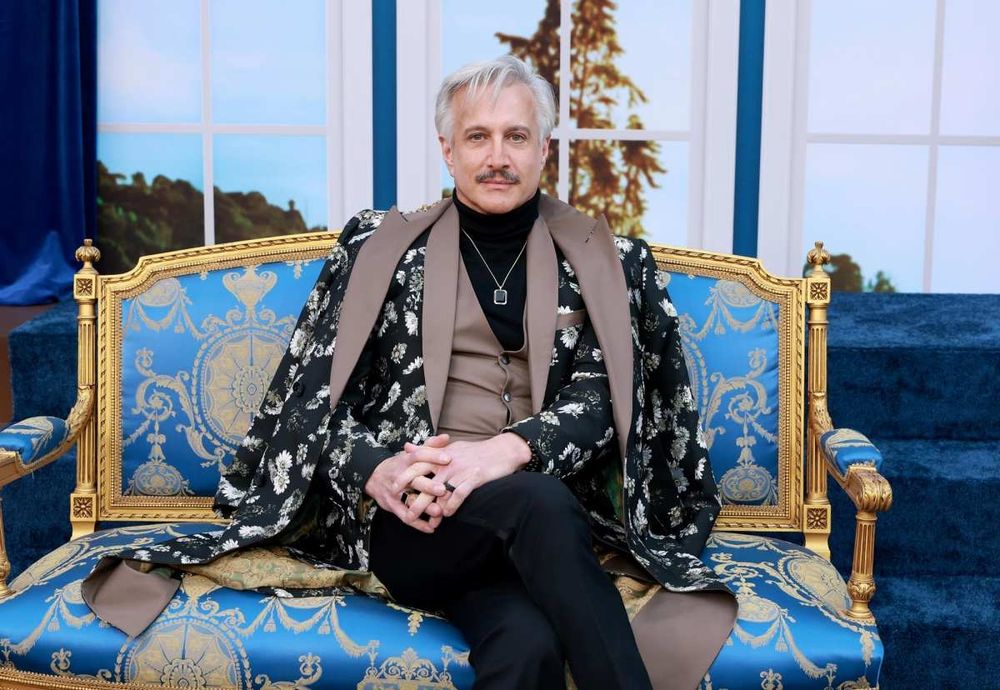 Bronson Pinchot as a gorgeous silver fox. He's wearing a black jacket of some sort with white flowers, and sitting on a fancy blue seat with gold designs