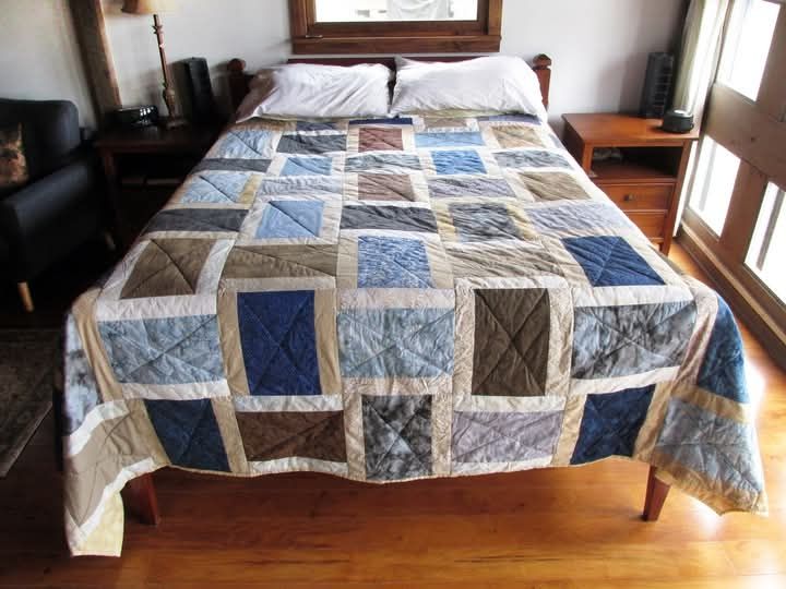 A handmade bed quilt with brown, gray, and blue fabrics on a bed beneath a window, made by Pam Geisel.