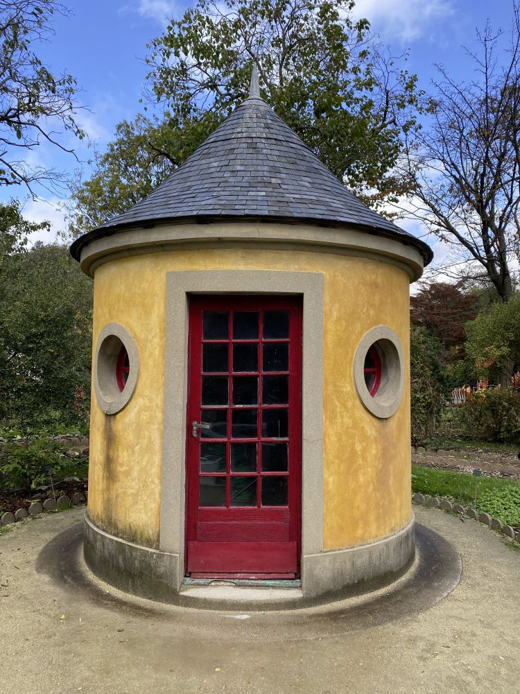 small round building in a Paris garden with a pointy slate roof, yellow walls, a red door and a small round window on each side of the door.