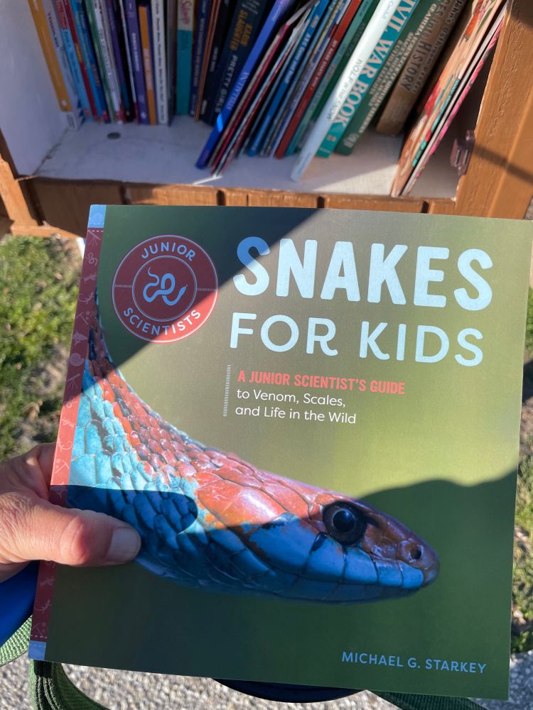 A copy of the book “Snakes for kids” is held up in front of a tiny neighborhood library. 
