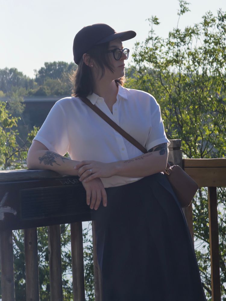 Joey leaning against a wooden railing in front of a river valley. They are wearing a navy blue ball cap, navy blue pleated hakama pants, a white short sleeved collared blouse, and brown leather purse. They are trying to look nonchalant like they weren’t having coughing fits immediately before and after this photo was taken. 
