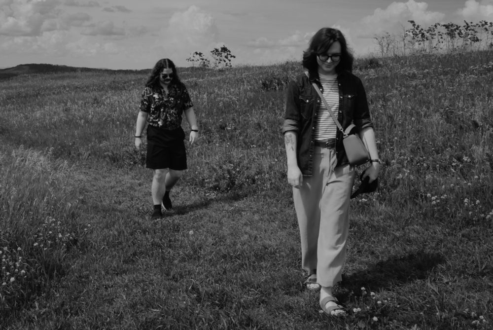 Black and white photo of Joey and Michael in a prairie. Joey is walking ahead of Michael. They both have their heads down. Joey is wearing a striped tee under a denim shirt with rolled up sleeves, with slacks and sandals. Michael is wearing a patterned button up and dark shorts. 