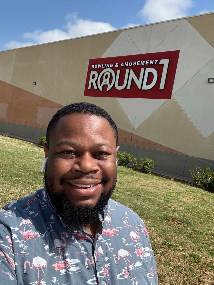 A selfie of Kelvin in front of a Round 1 Building Sign. He is wearing a flamingo based shirt and smiling. 