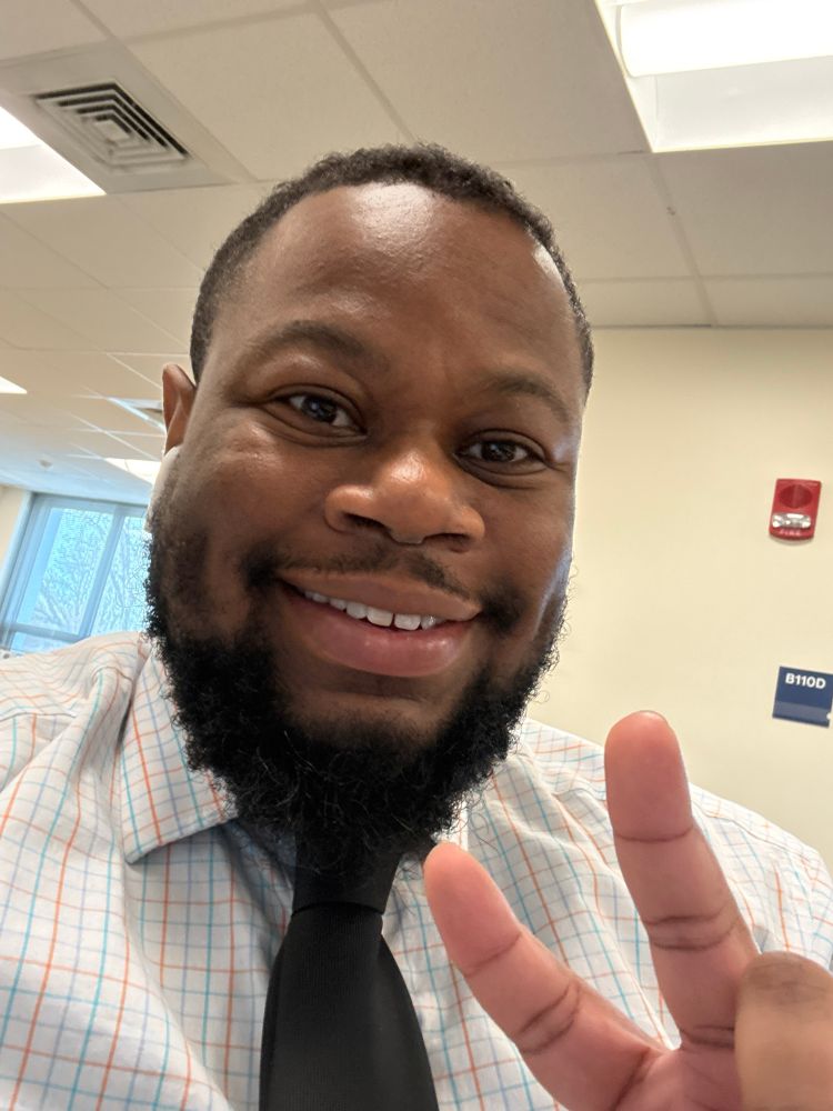 A selfie of Kelvin smiling. He is wearing a striped collared shirt with a dark gray tie and is holding two fingers in a peace sign. 