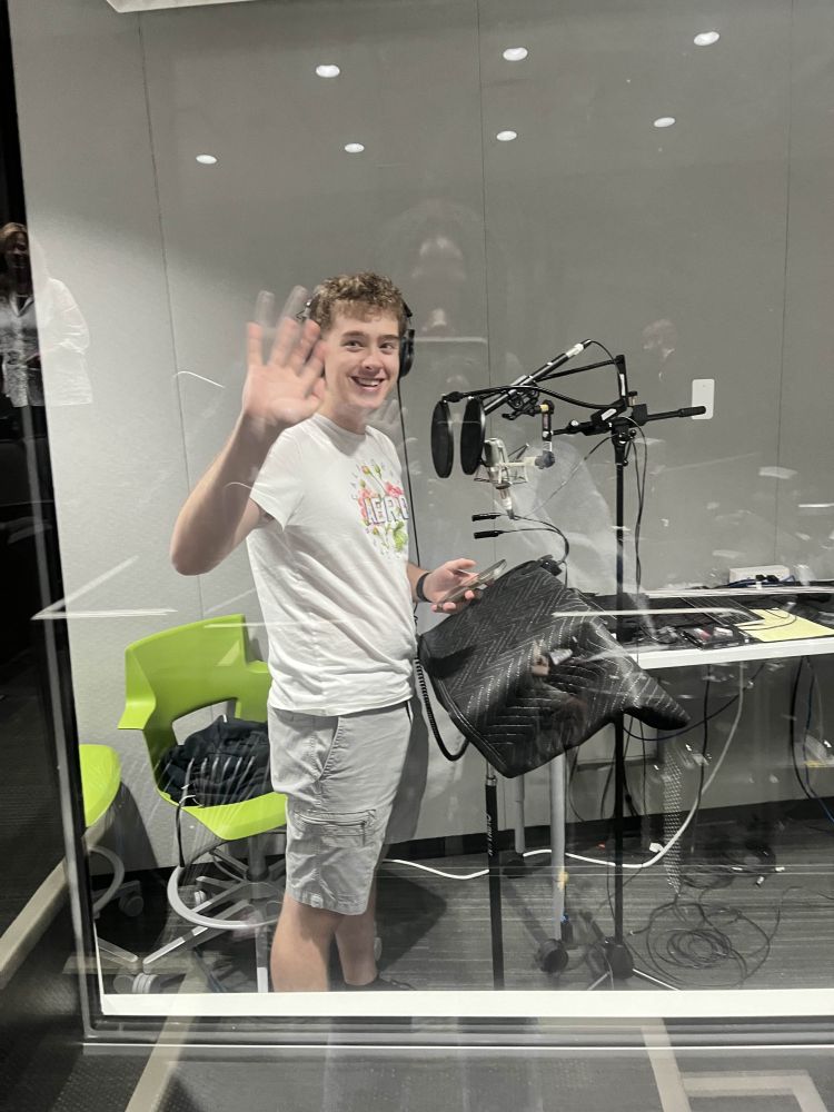 Me, moonlighting as a voice actor! I am standing in a voice acting booth, wearing a white Aero t-shirt, smiling and waving while a friend takes a photo of me from the outside of the booth.