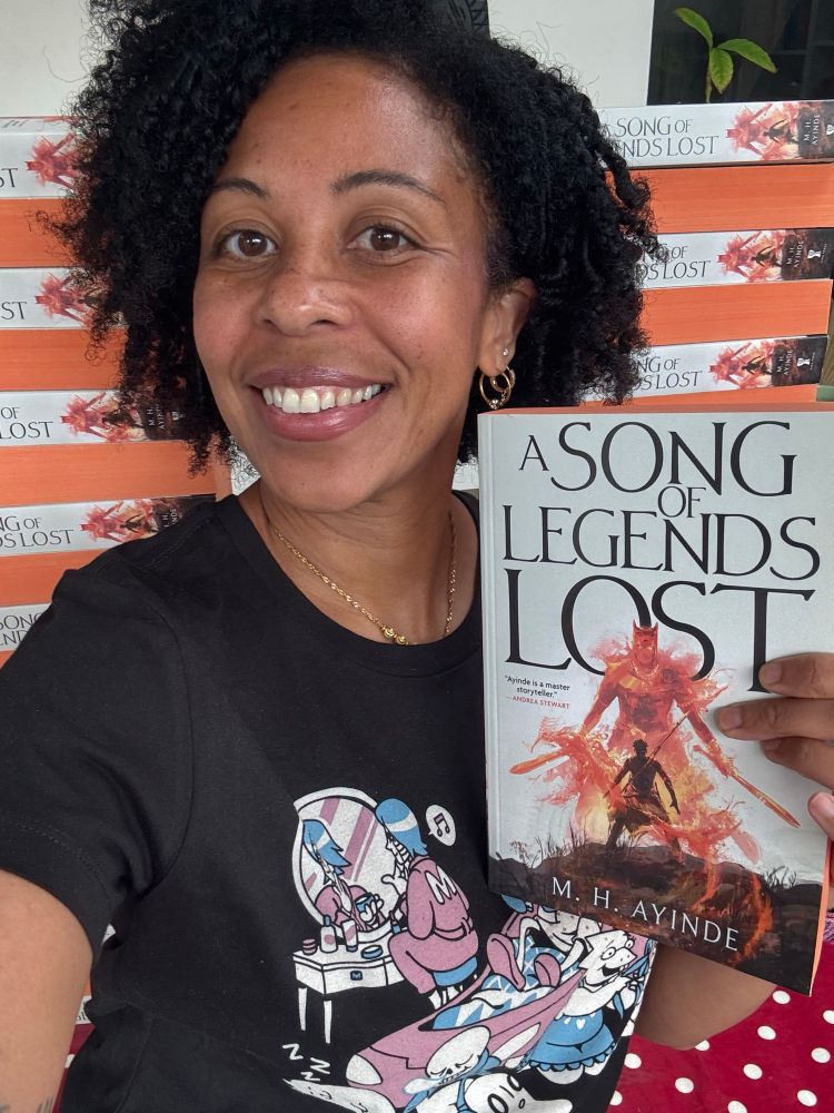 Modupe holding up a paperback copy of A SONG OF LEGENDS LOST. She’s wearing an Undertale t-shirt