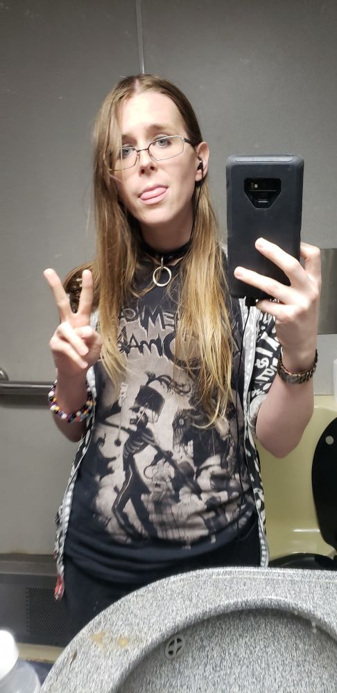 bunni (it/she) in the bathroom on a train. it's wearing a collar, a My Chemical Romance shirt with the Black Parade album artwork, and a Paramore hoodie that says "RIOT!" all over