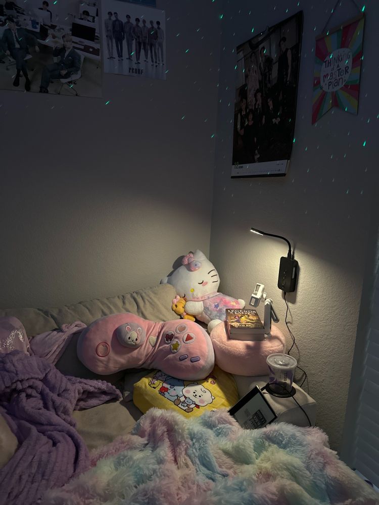 a chaise lounger covered in pastel pillows and blankets with books resting on the arm of the chair and a book lamp affixed to the wall.