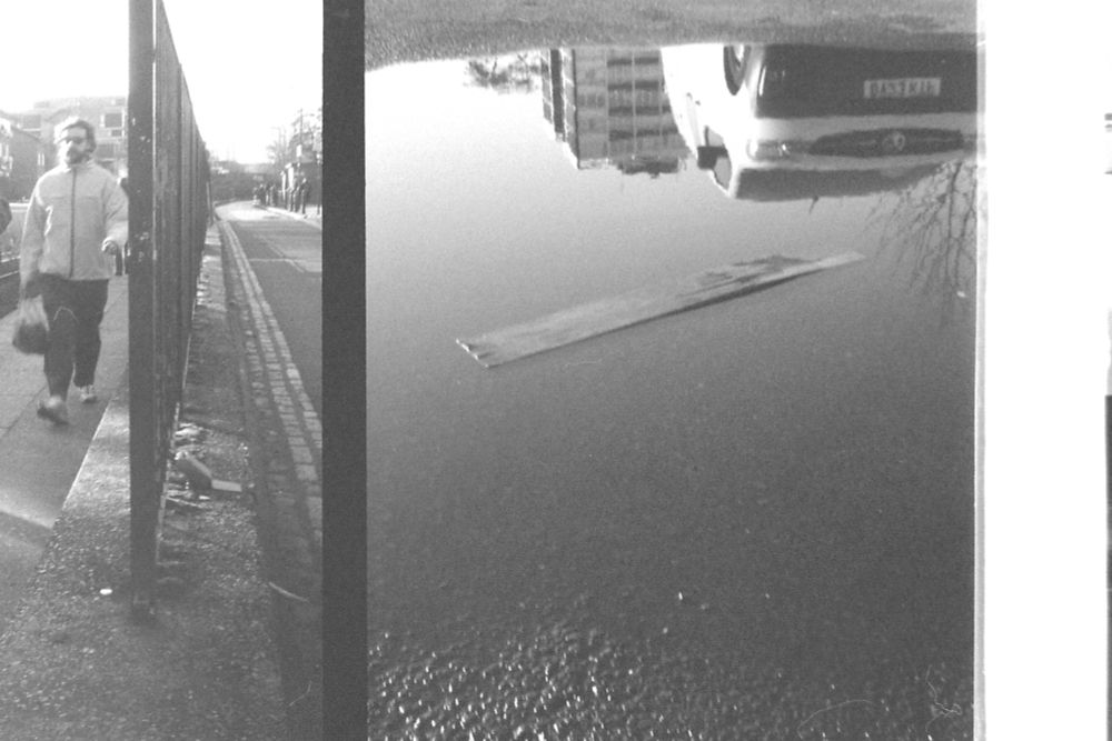 Ilford FP4+ 35mm detail across two half frame images taken on an Olympus Pen EE2 with no red flag, so assuming no selenium metering. Showing in the left frame a man walking towards camera along the Regents Canal, and in the right frame a reflection in a puddle of a van with a block of flats in the background.