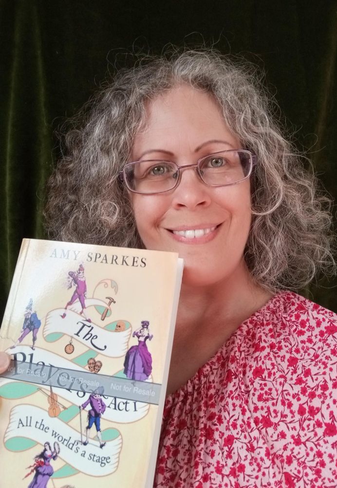 A smiling Amy Sparkes stands in front of a green curtain, holding a copy of her new historical fiction book: THE PLAYERS ACT 1: ALL THE WORLD'S A STAGE. 
