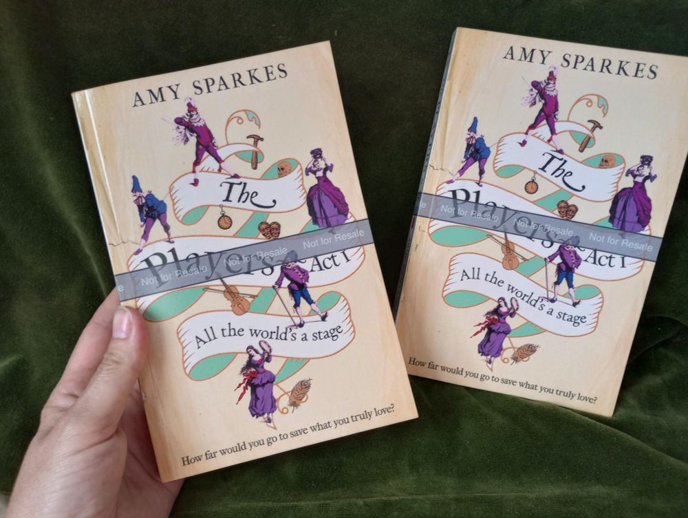 Two copies of THE PLAYERS ACT 1: ALL THE WORLD'S A STAGE rest against a green curtain.  The cover has a parchment-coloured background with the title spiralling down on a pale ribbon. Five commedia dell'arte character in purple are dotted around. There are various objects such as a hammer, a skull, a pocket-watch, the theatre masks and a fiddle visible on the cover. A grey proof copy ribbon runs across the cover saying 'Not for resale'.


