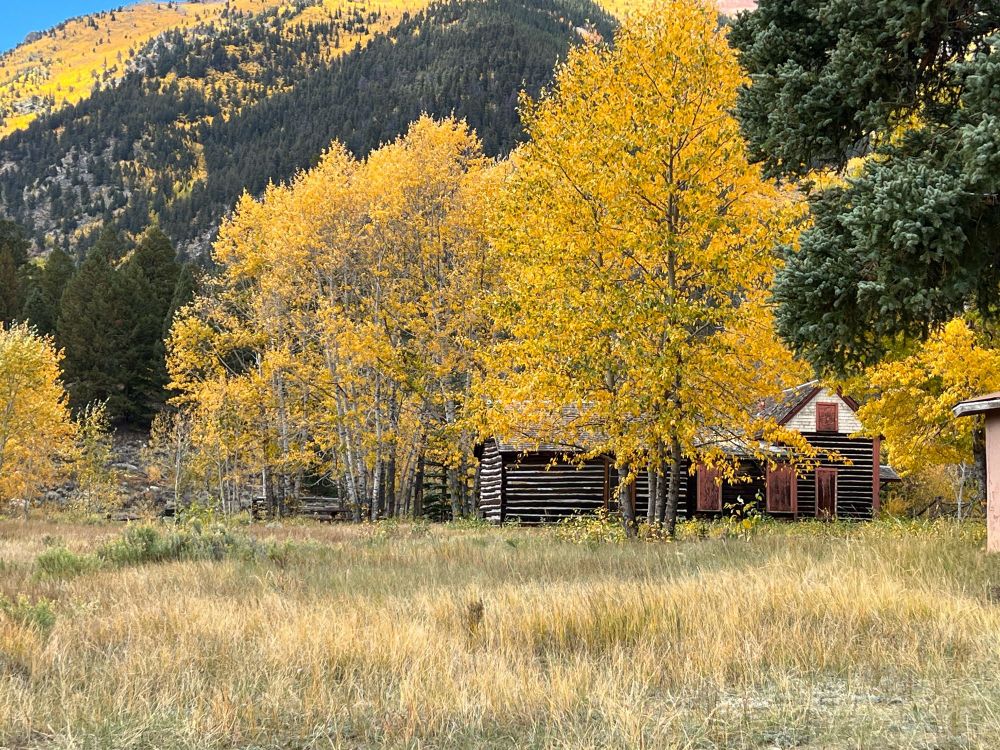 An old house stands in front of fall mountains, yellow and green leaves on the trees.
